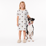 Girl wearing a white nightgown with dog print standing next to a dog on a white background