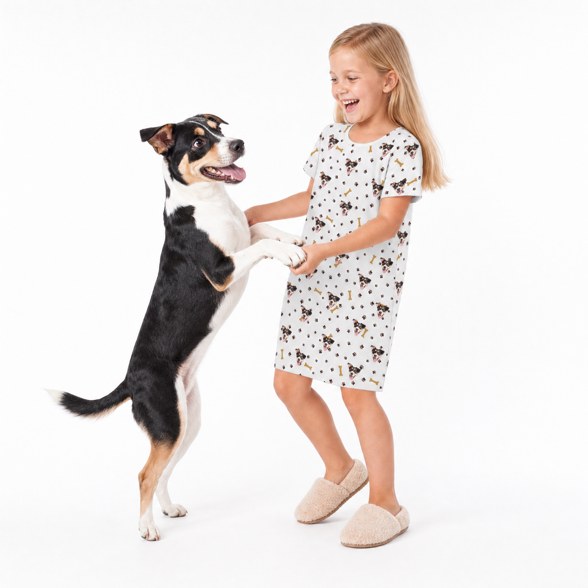 Young girl wearing a white paws and bones print sleep shirt while standing in slippers, smiling and holding the front paws of a happy black, white, and tan dog standing on its hind legs.