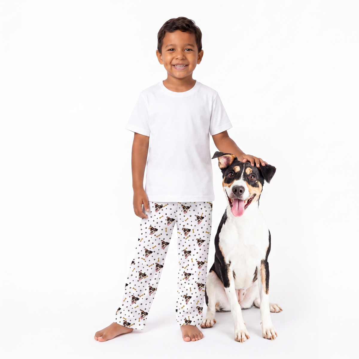 A young boy wearing a white t-shirt and paw and bones pajama pants with a black and brown dog print stands next to a black and white dog on a solid white background.
