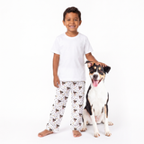 A young boy wearing a white t-shirt and paw and bones pajama pants with a black and brown dog print stands next to a black and white dog on a solid white background.