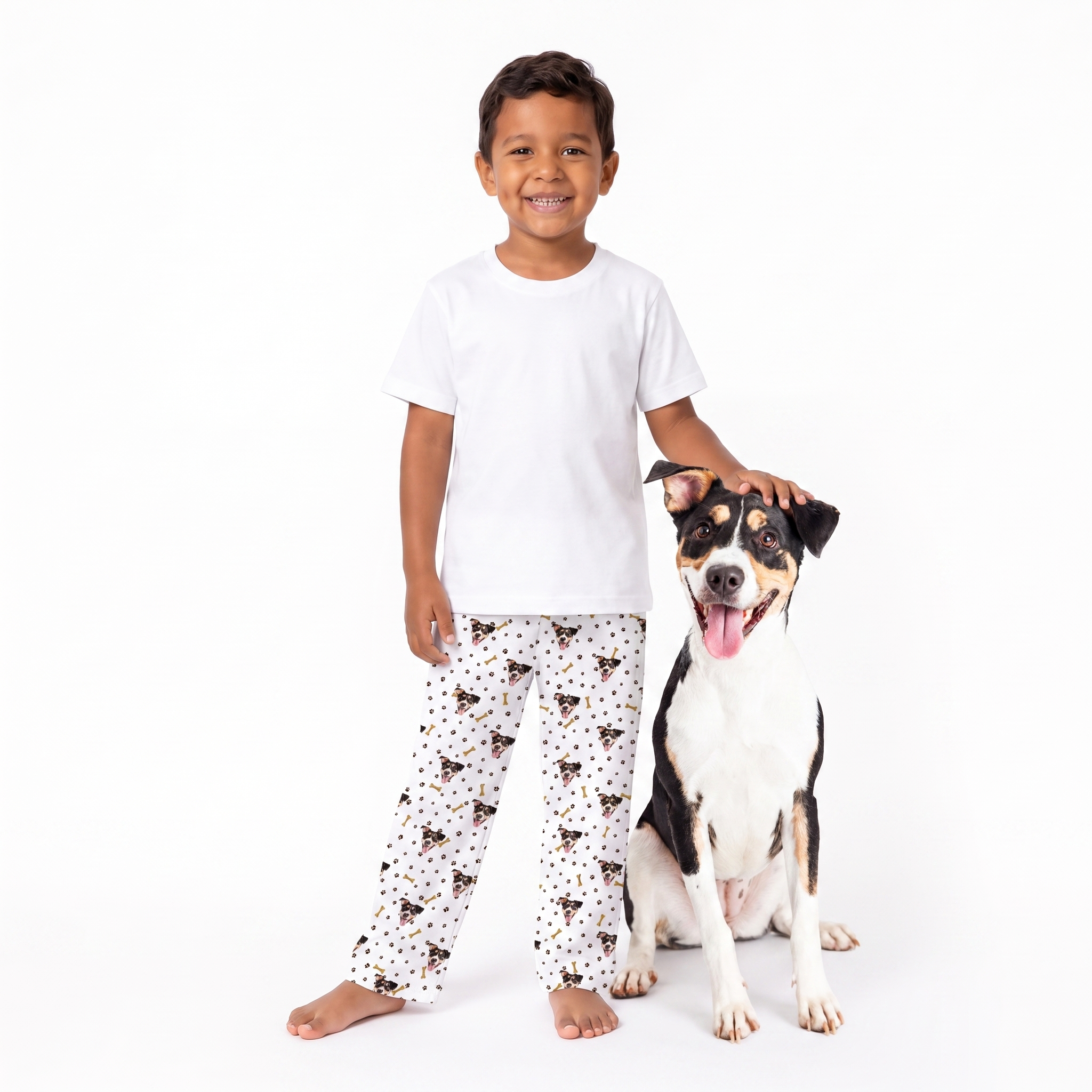 A young boy wearing a white t-shirt and paw and bones pajama pants with a black and brown dog print stands next to a black and white dog on a solid white background.