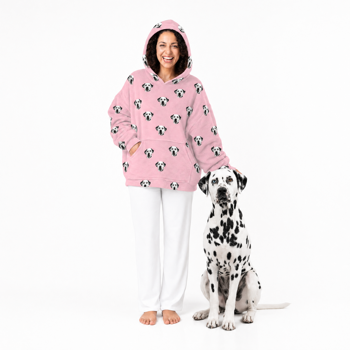 Woman wearing a pink blanket hoodie with dog patterns and white pants, standing next to a Dalmatian dog on a white background.