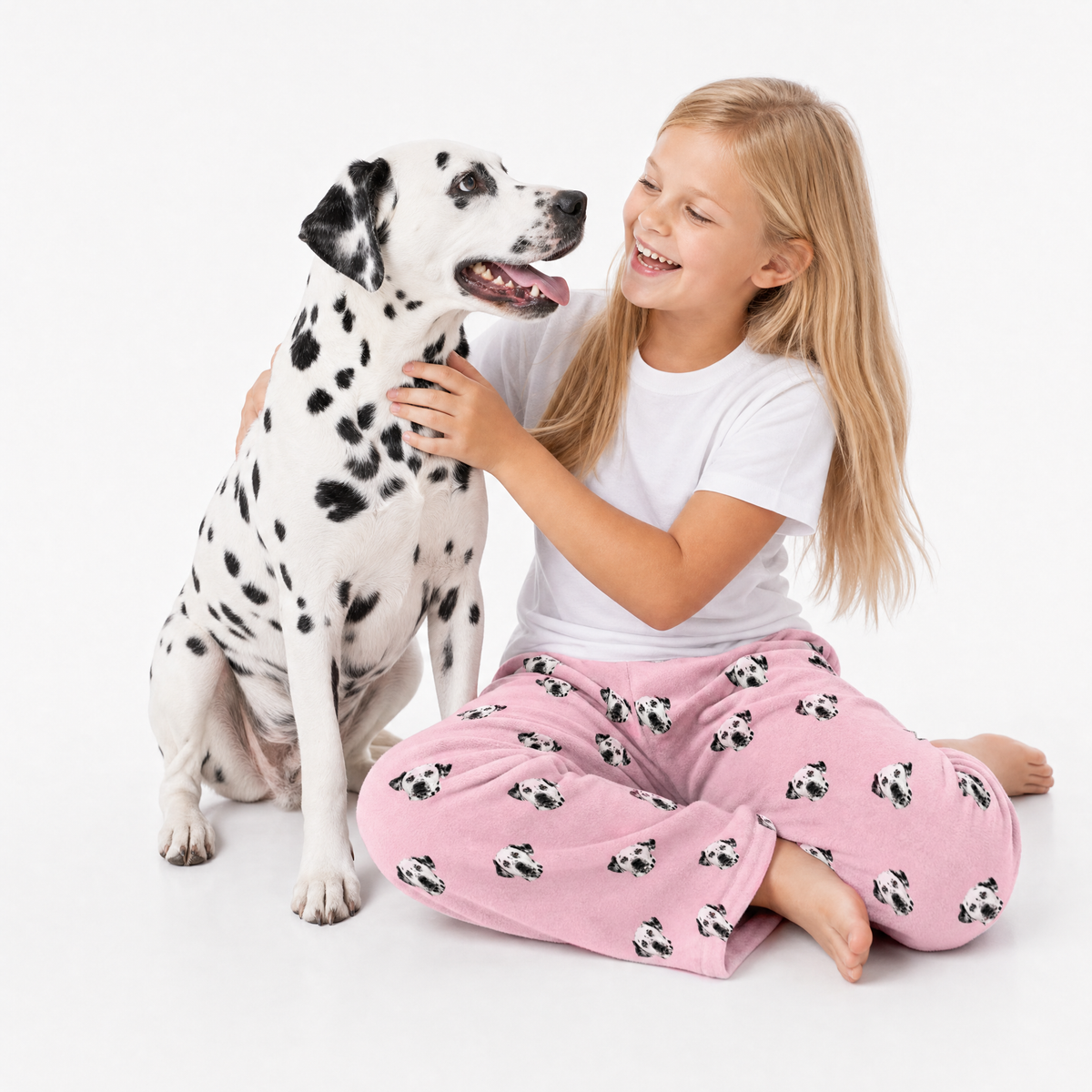 Young girl in pink pajama pants with dog pattern sitting next to a Dalmatian dog on a white background