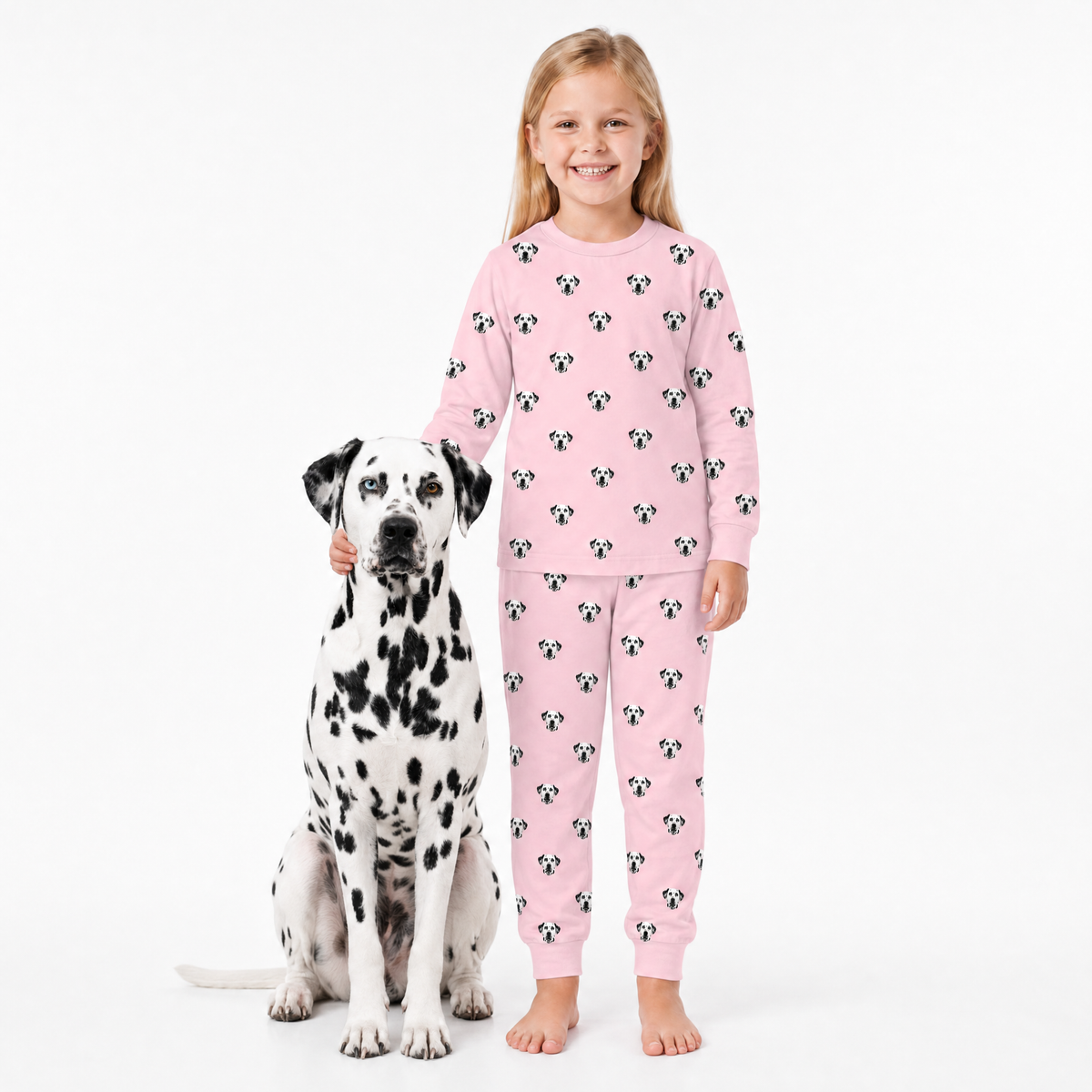 Child wearing pink jogger pajamas with dog pattern standing next to a Dalmatian dog on a white background