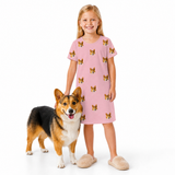 Young girl in a pink nightgown with dog pattern standing next to a corgi on a white background