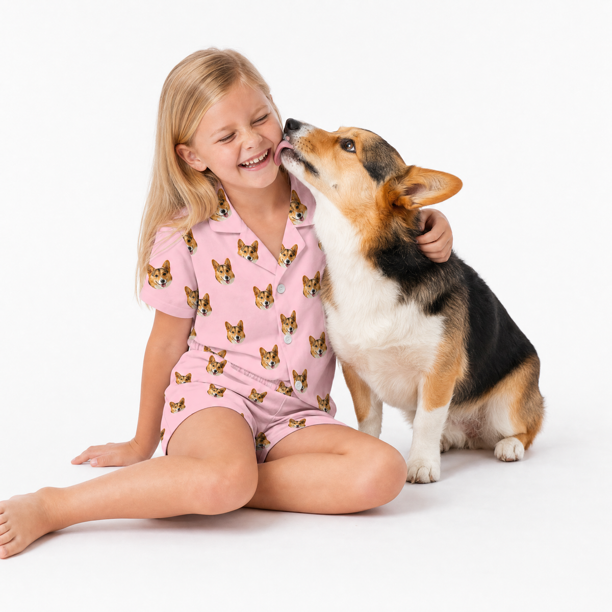 Young girl in pink pajamas with dog pattern sitting next to a corgi on a white background