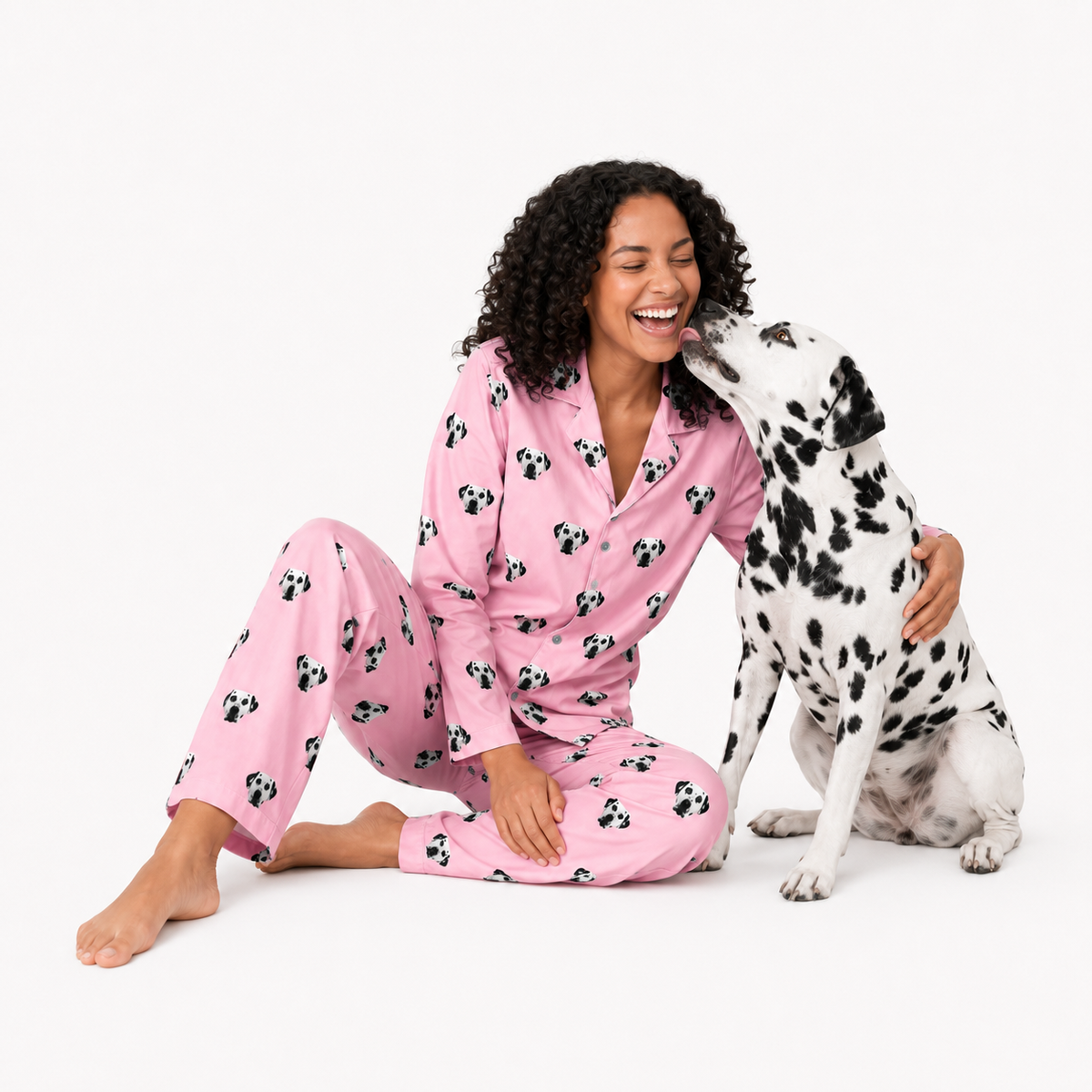 Woman in pink pajamas with dog pattern sitting next to a Dalmatian dog on a white background