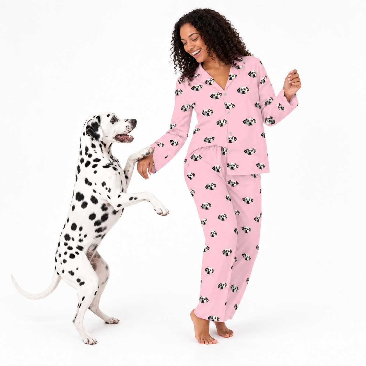 Woman in pink pajamas with dog pattern standing next to a Dalmatian dog on a white background