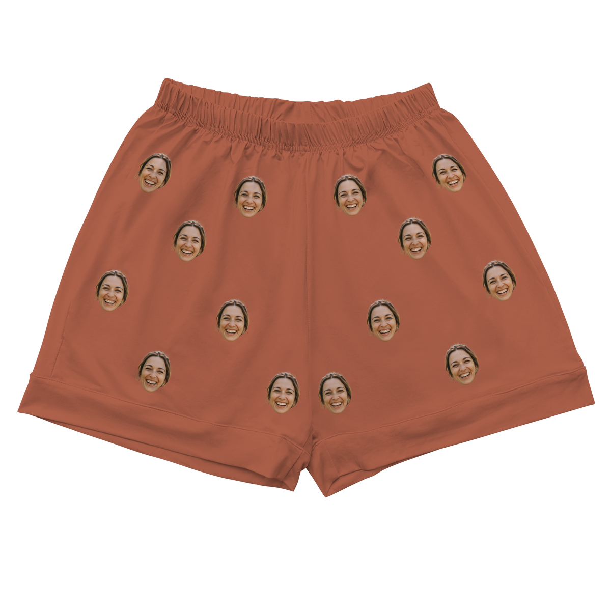 A pair of athletic or pajama shorts with a gathered, elastic waistband, made from a solid burnt orange or cocoa-colored fabric. Repeated headshots of a smiling woman's face are evenly spaced and superimposed across the fabric