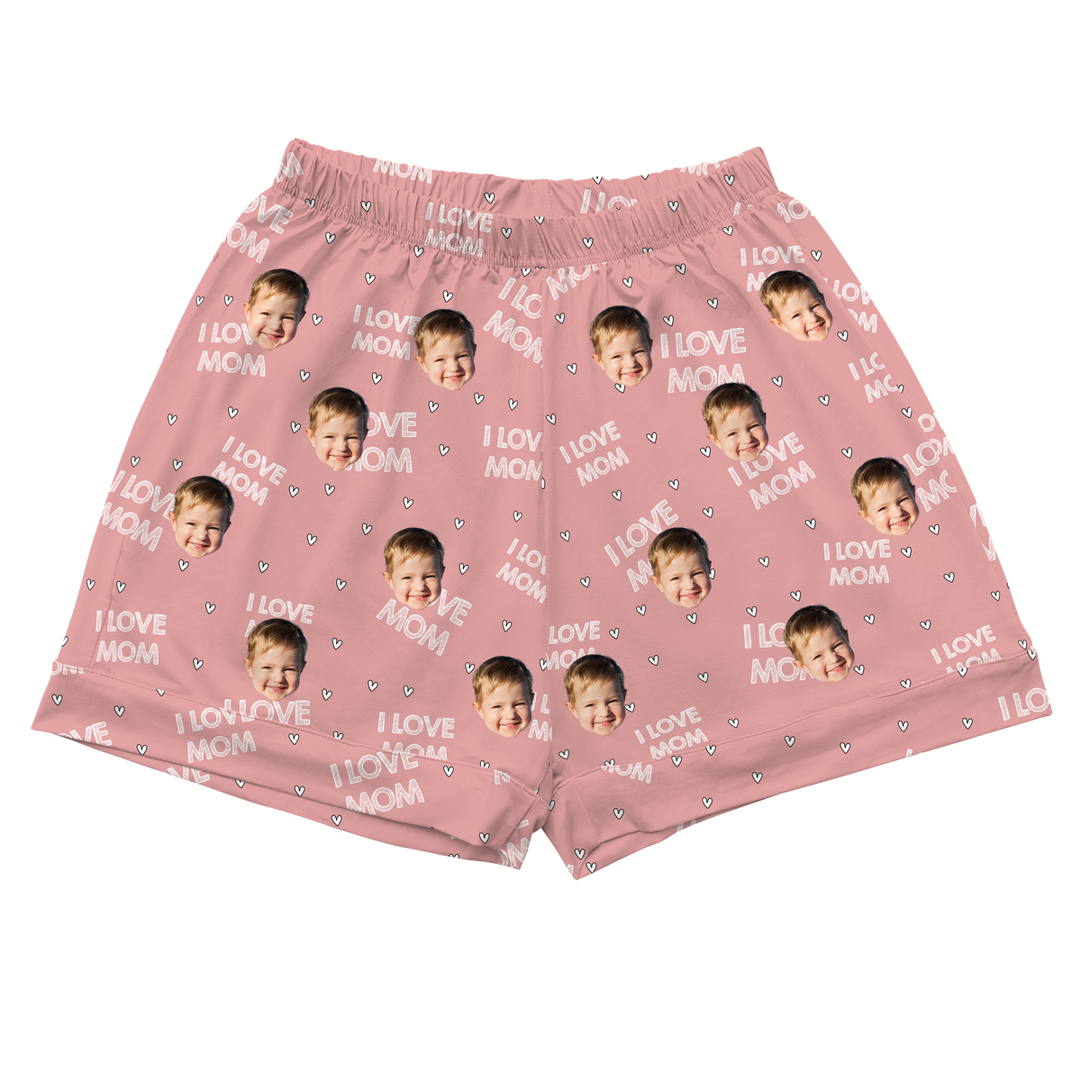 A pair of athletic or pajama shorts with a gathered, elastic waistband, made from a light pink fabric. The fabric is patterned with the repeating white text "I LOVE MOM" and small white heart outlines. Repeated headshots of a baby's smiling face are evenly spaced and superimposed across the fabric. 