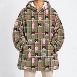 A person wearing an oversized green plaid hoodie with a repeating print of Gatsby cats in pink outfits and small pink and blue logo patches, photographed against a plain light gray studio background.