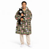 A man wearing a green plaid hooded robe with a black cat print pattern, holding a black and white cat, standing against a white background.