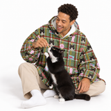 A man sitting on a white studio floor wearing a green plaid hoodie with a black cat print pattern, interacting with a black and white cat. The hoodie features a green and red plaid base with cartoon black cats, pink bows, and small colorful patches.