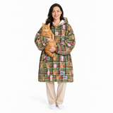 A woman stands against a white studio background wearing a green hooded sweatshirt with a colorful cat-themed plaid pattern and holding an orange tabby cat. She is also wearing beige pants and white socks.