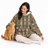 A woman sits cross-legged on a white studio floor wearing a green plaid hooded sweatshirt covered in a cat print pattern, holding an orange tabby cat. She is wearing beige sweatpants and white socks.