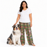 A woman stands barefoot next to a fluffy Australian Shepherd dog, wearing green plaid pajama pants with a dog-themed print and a plain white t-shirt, against a solid white background.
