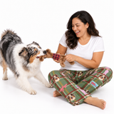 A woman sitting on a white studio floor plays tug-of-war with a fluffy Australian Shepherd dog wearing green plaid pajamas with dog faces. The woman holds a brown plush toy with a red plaid pattern.