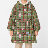 A person wearing a green plaid hooded sweatshirt with an all-over print of dog faces, pink figures, and small blue-and-white logo patches, photographed against a plain light gray studio background.