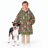 A smiling boy stands with a black, white, and tan dog against a plain white studio background, wearing a long green plaid hooded blanket or coat covered in dog-themed illustrations.