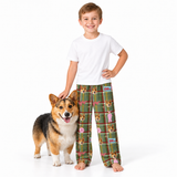A young boy wearing a white t-shirt and green plaid pajama pants with a dog print stands next to a corgi on a white studio background.