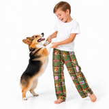 A young boy wearing green plaid pajama pants with a dog print stands barefoot on a white studio background, playfully interacting with a corgi dog that is standing on its hind legs to reach his hand.