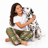 A young girl with long brown hair sits on a white floor hugging a Dalmatian dog. She is wearing a white t-shirt and green plaid pajama pants with dog face prints and pink bows.