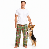 A man wearing green plaid pajama pants with a dog print stands barefoot next to a corgi on a white studio background. The pajama pants feature a green plaid pattern with pink bows, red stripes, and cartoon dog faces.
