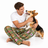 A man sitting cross-legged on a white studio floor, hugging a Corgi mix dog. He is wearing green plaid pajama pants with a cartoon dog pattern and a white t-shirt.