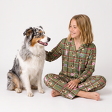 A girl wearing green plaid pajamas with a dog print sits cross-legged on a white studio floor next to an Australian Shepherd dog.