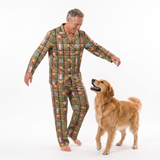A man wearing a green plaid pajama set with a dog print pattern stands barefoot on a white studio background, smiling as he looks at a golden retriever dog.