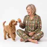 A woman wearing green plaid pajamas with a dog print sits cross-legged on a white studio floor, holding an orange tennis ball for a small brown poodle.