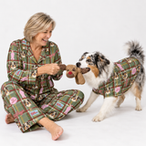A woman and a dog wearing matching green plaid pajamas with dog illustrations, sitting on a white studio floor and playing with a brown plush toy.