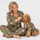 A woman and her small dog sit together on a white studio background, both wearing matching green plaid pajamas with cartoon dog faces and pink bows. The woman is smiling and offering a treat to the dog.