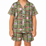 A child wearing a matching short-sleeve button-front pajama top and shorts set in an olive green plaid fabric with dog face prints, pink script text, and small blue and pink logo patches, photographed against a plain white background.
