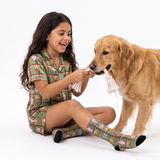A young girl with long dark hair sits on a white studio floor smiling while playing with a golden retriever dog. The girl is wearing a green plaid pajama set with a cartoon dog pattern, and the dog is holding a white rope tug toy.
