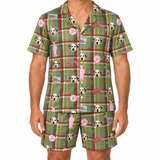 A man wearing a matching short-sleeve button-front pajama shirt and shorts set with a green plaid pattern, featuring Dalmatian dog faces, pink speech bubbles, and small cartoon graphics on a white background.