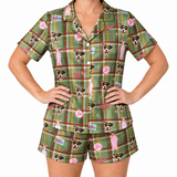 A woman modeling a matching short-sleeve button-front pajama top and shorts set in green plaid with dog face prints, pink figures, and small logo patches, photographed against a plain white background.