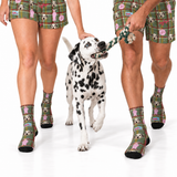 A Dalmatian dog walks on a white studio background while being held on a rope toy by two people wearing matching green plaid socks and shorts with dog-themed prints. The socks and shorts feature dog faces, beds, and small pink accents.