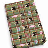 A wrapped gift box with a green plaid holiday pattern featuring black-and-white cat faces, pink teddy bears, and small blue signs, tied with twine on a white background.