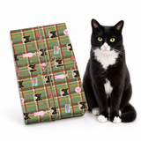 A black and white cat sits next to a wrapped gift box featuring a green plaid pattern with cat faces and pink hearts, on a clean white background.
