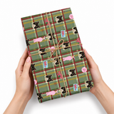 A person's hands holding a wrapped gift box featuring a green plaid pattern with black cats, pink bunnies, and small blue gift tags, tied with twine on a white background.