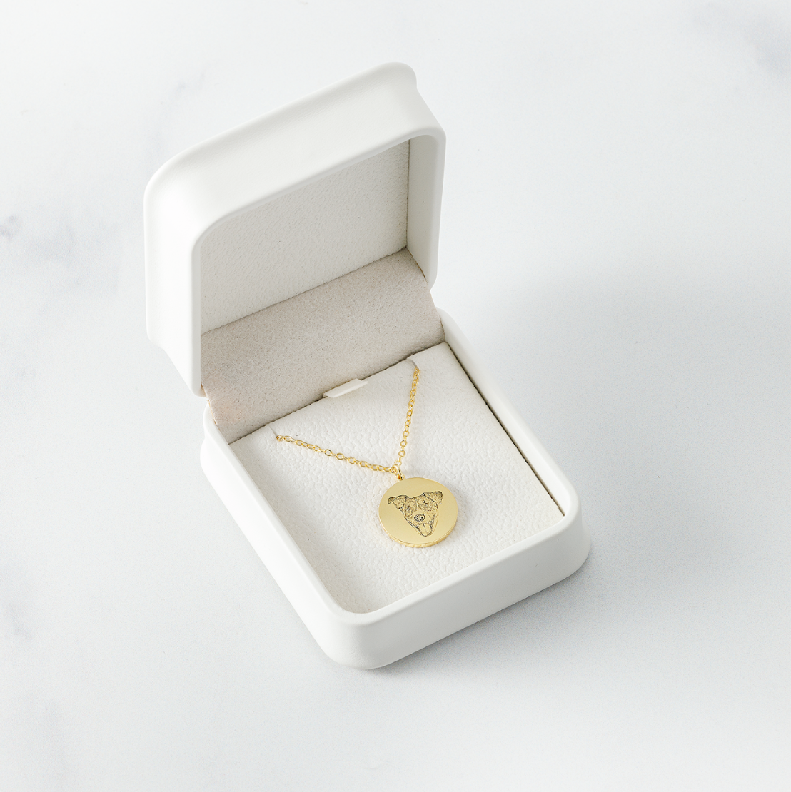 Gold dog-portrait pendant in a white velvet jewelry box on a marble surface.