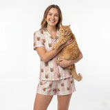 Woman wearing a pajama set with cat designs, holding an orange cat on a white background [TerracottaStripes]