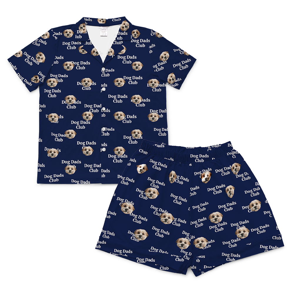 A matching short-sleeve pajama set (top and shorts) in a dark navy blue fabric. The entire set is covered in a repeating pattern featuring alternating elements: the phrase "Dog Dads Club" in white text, and headshots of a dog.