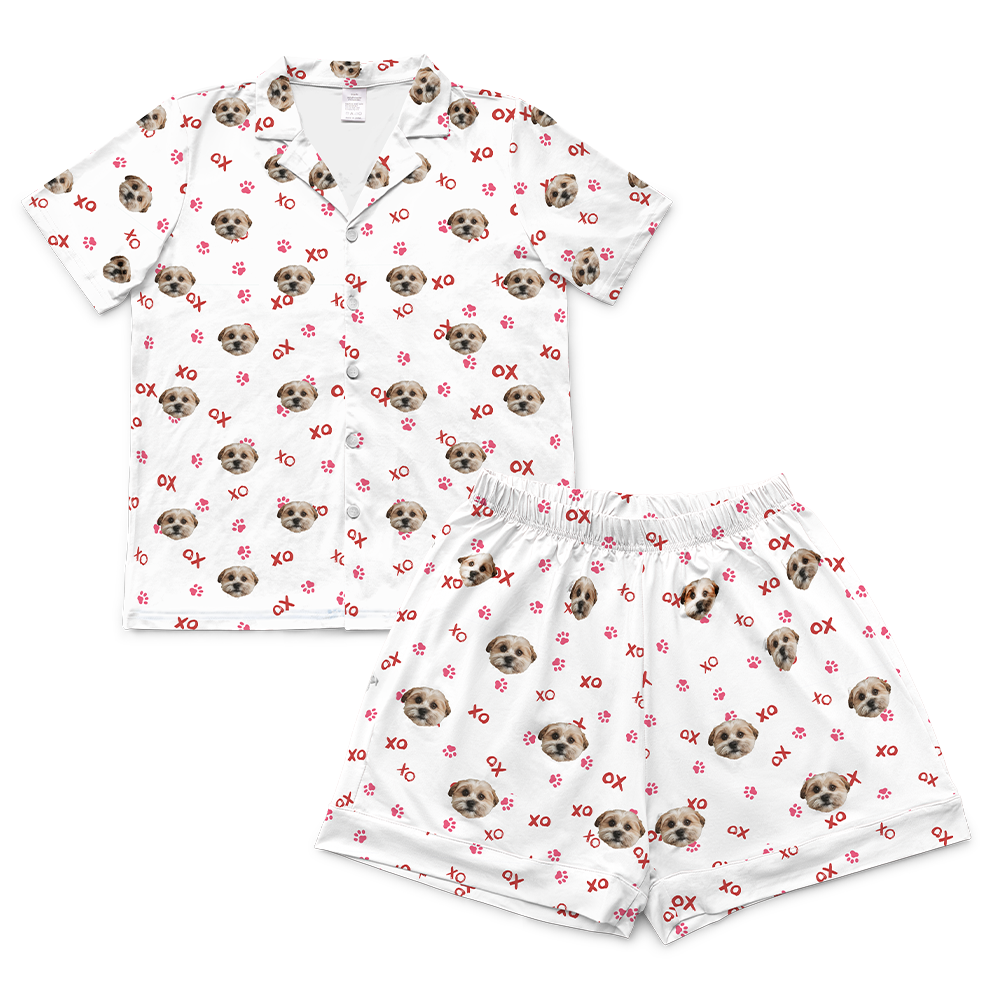White short-sleeve pajama set featuring a button-down top and matching shorts, both printed with a single dog’s face, pink paw prints, and red “XO” lettering in an all-over pattern.