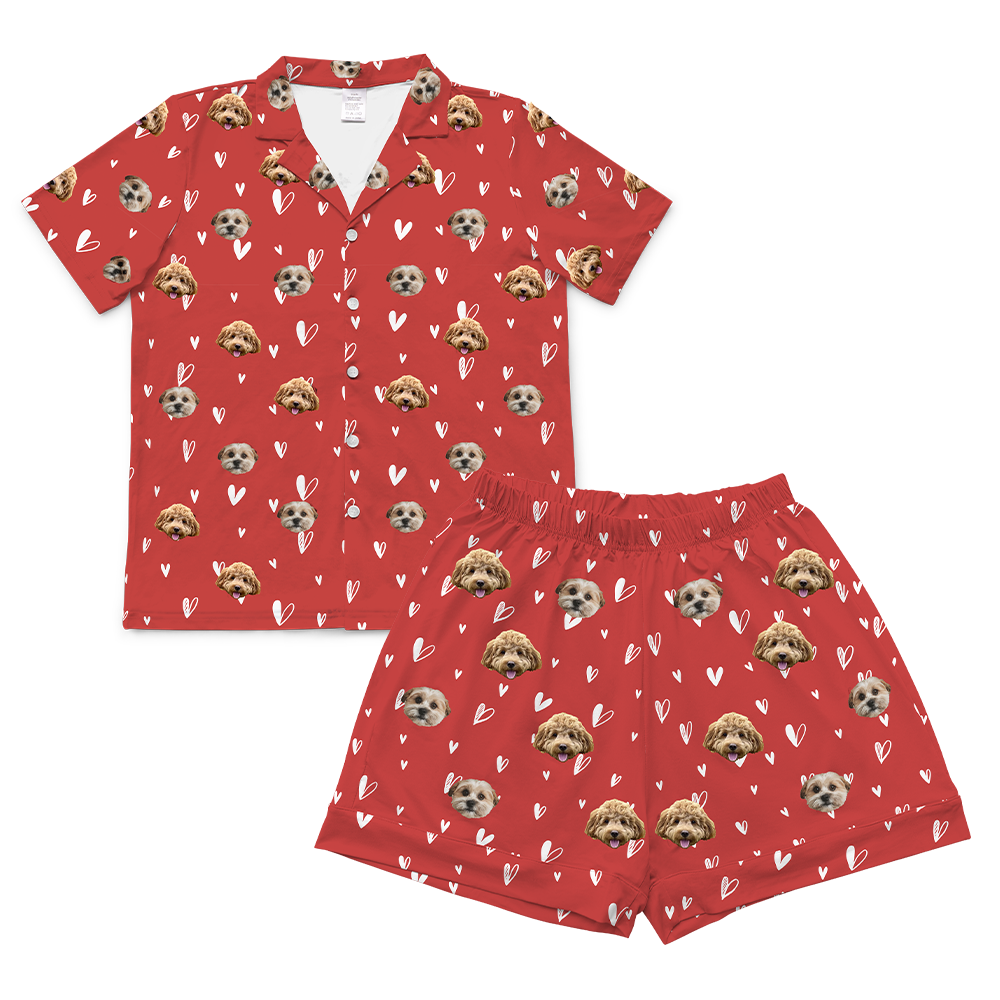 A single short-sleeve, button-down pajama shirt in a vibrant red color. The fabric has an all-over pattern of scattered white line-art and solid hearts. Overlaid on this pattern are repeating, cropped dog faces.