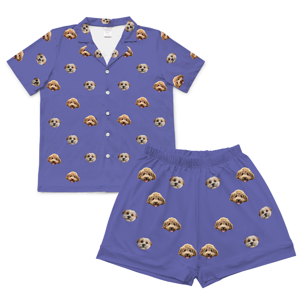 A short-sleeve button-down pajama top in a grape purple color, laid flat on a light gray background. The top has a collar and features a scattered, repeating pattern of two different dog faces.