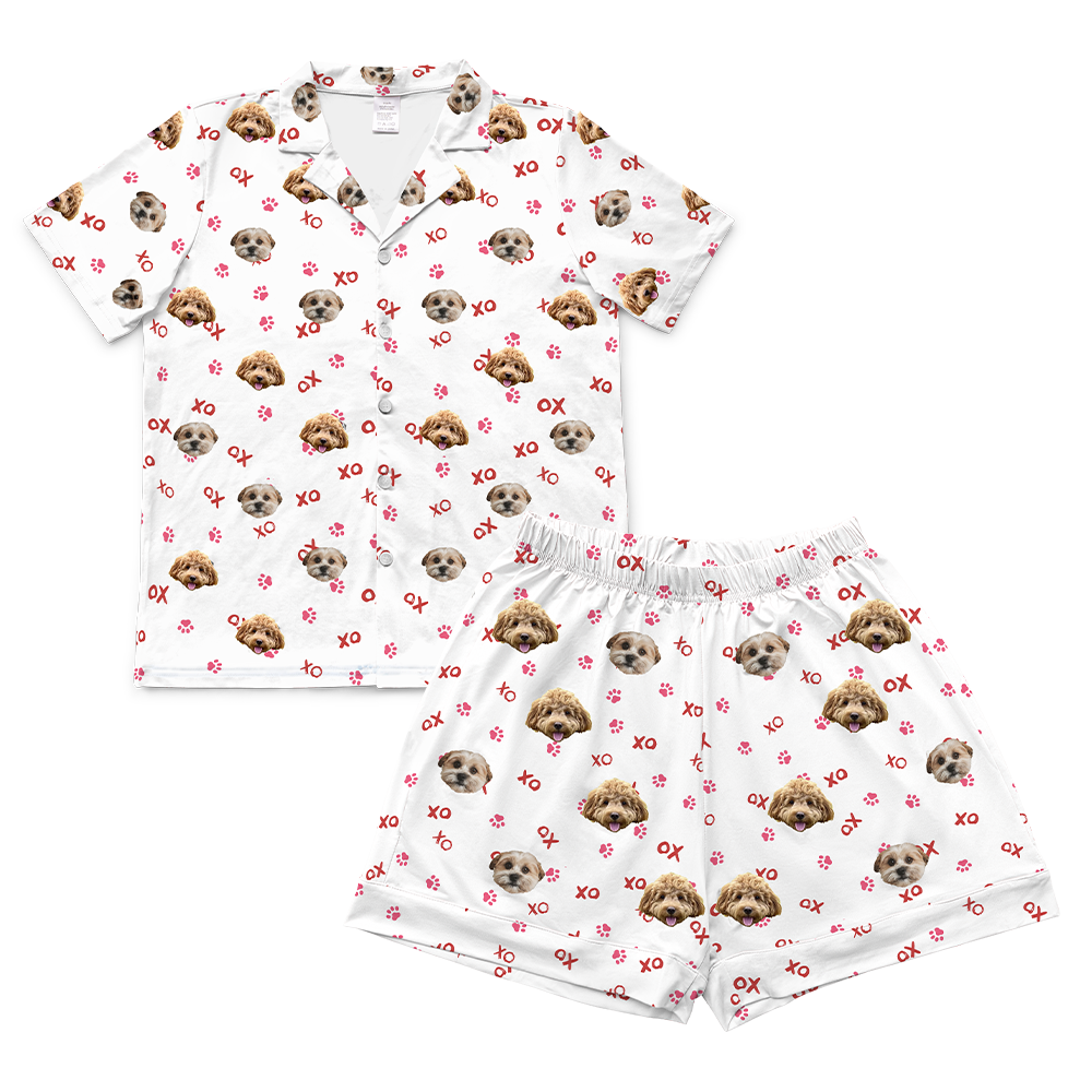 A matching pajama set featuring a short-sleeved, button-down top with a collar and a pair of shorts with an elastic waistband. Both pieces are white and display a pattern of two different dogs' faces, along with scattered red "XO" text and pink red prints.