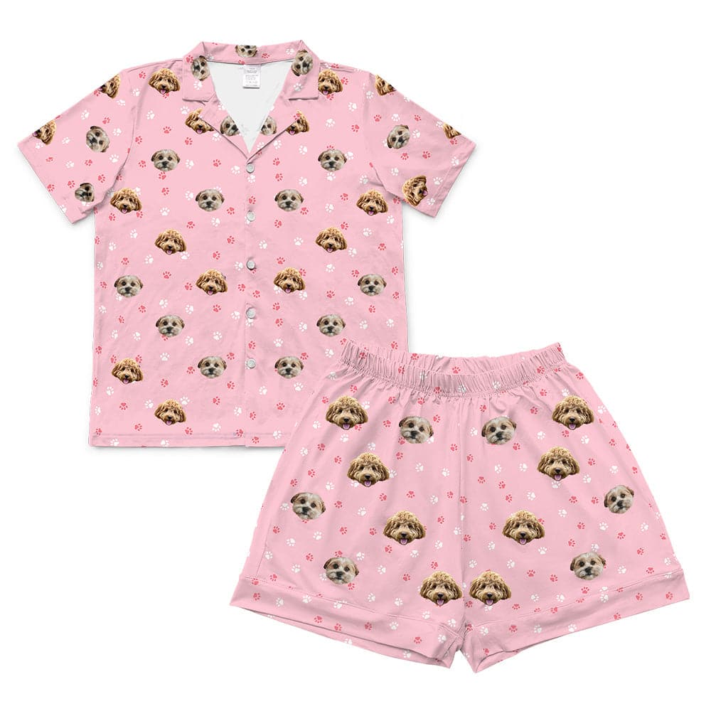 A two-piece pajama set, consisting of a short-sleeved, button-down top and matching elastic-waist shorts. Both pieces are light pink and feature a repeated pattern of two different dog faces.