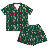 A two-piece matching set featuring a short-sleeve button-up scrub top and shorts in dark green with an all-over Christmas-themed print of Dalmatian dogs, Santa Claus, Christmas trees, and holiday decorations, displayed on a clean white background.